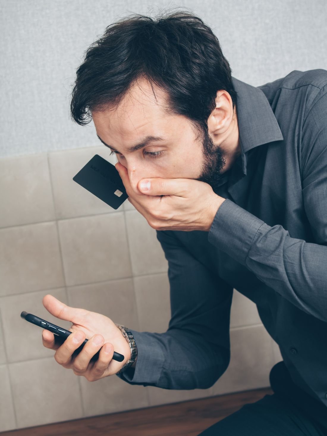 Hombre preocupado revisando su teléfono mientras sostiene una tarjeta bancaria