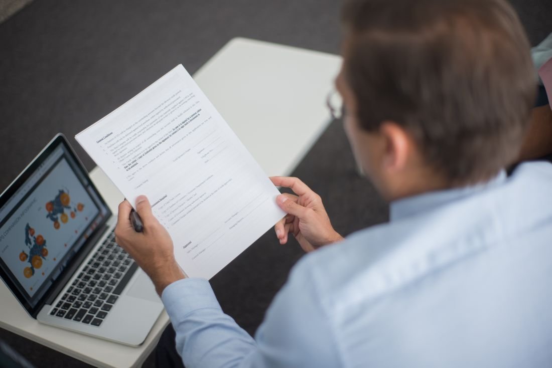 Hombre revisando un documento impreso frente a un computador portátil sobre una mesa
