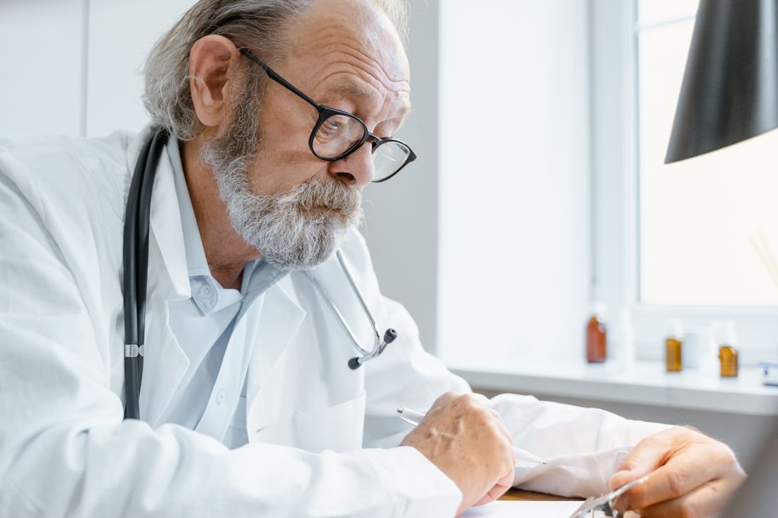 Médico con bata blanca y estetoscopio escribiendo en su consultorio